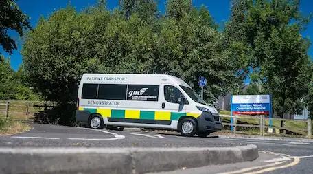 White GM Coachwork patient transport demonstrator van with green and yellow reflective chevrons parked outside Newton Abbot Community Hospital.