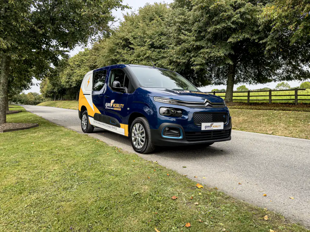 Front three-quarter view of a dark blue Citroën ë-Berlingo electric van with white and yellow GM Mobility branding, parked on a paved lane alongside a grassy tree-lined tree verge.